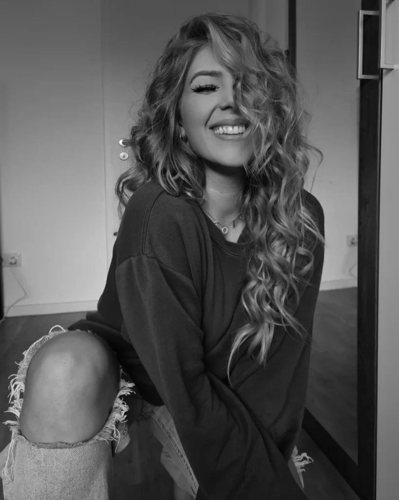 Hailee Summer sits in casual attire in front of a mirror, smiling naturally.