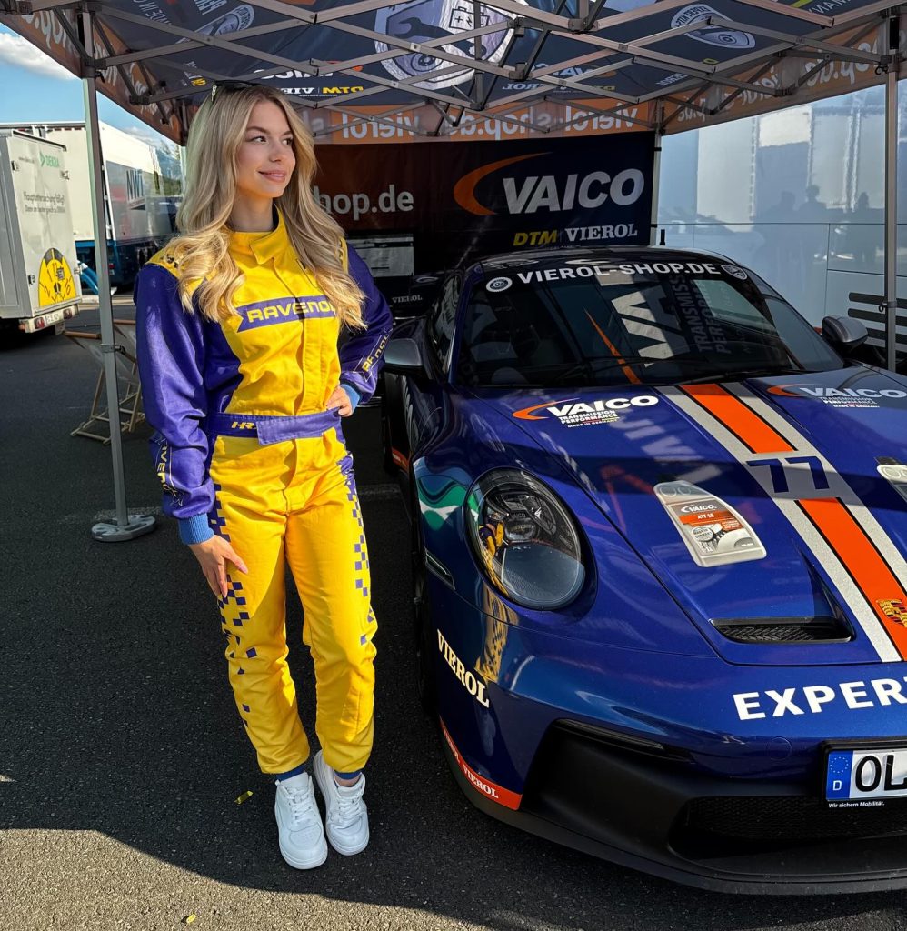 Blonde woman in a yellow and purple racing suit stands in front of a blue racing car with orange details under a Vierol tent, ready for a motorsport event.