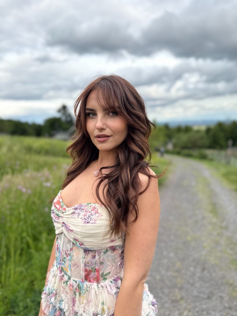 Mary A Ann steht auf einem Feldweg, trägt ein helles, florales Sommerkleid und blickt mit einem sanften Lächeln in die Kamera, umgeben von grüner Landschaft und bewölktem Himmel.