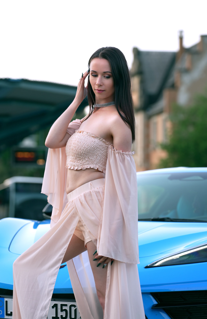 Ronya Rabe poses in an elegant, rose-colored outfit in front of a blue sports car – sensual, modern, and stylish.