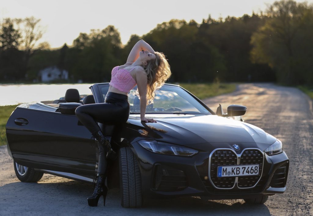 Mika Nox leaning against a black BMW convertible at sunset wearing a pink crop top and black leather pants
