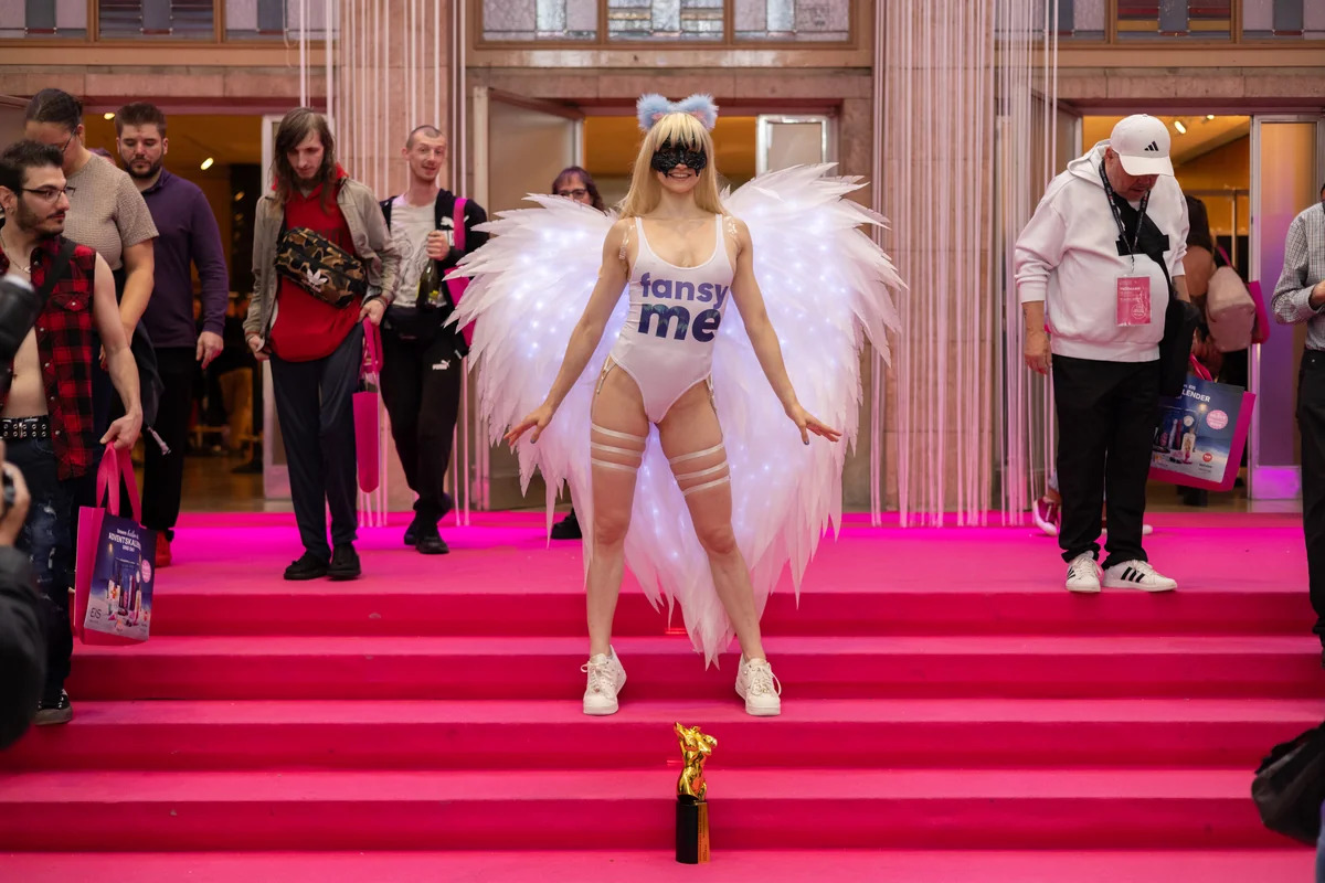 Ana Lingus in a white FansyMe bodysuit with illuminated angel wings and a mask on the pink carpet at Venus Berlin 2025