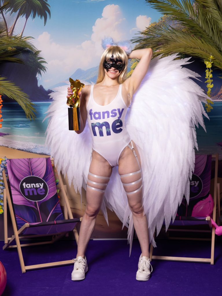 Ana Lingus in a white FansyMe bodysuit with angel wings and a mask, holding a trophy at the FansyMe booth at Venus Berlin 2025