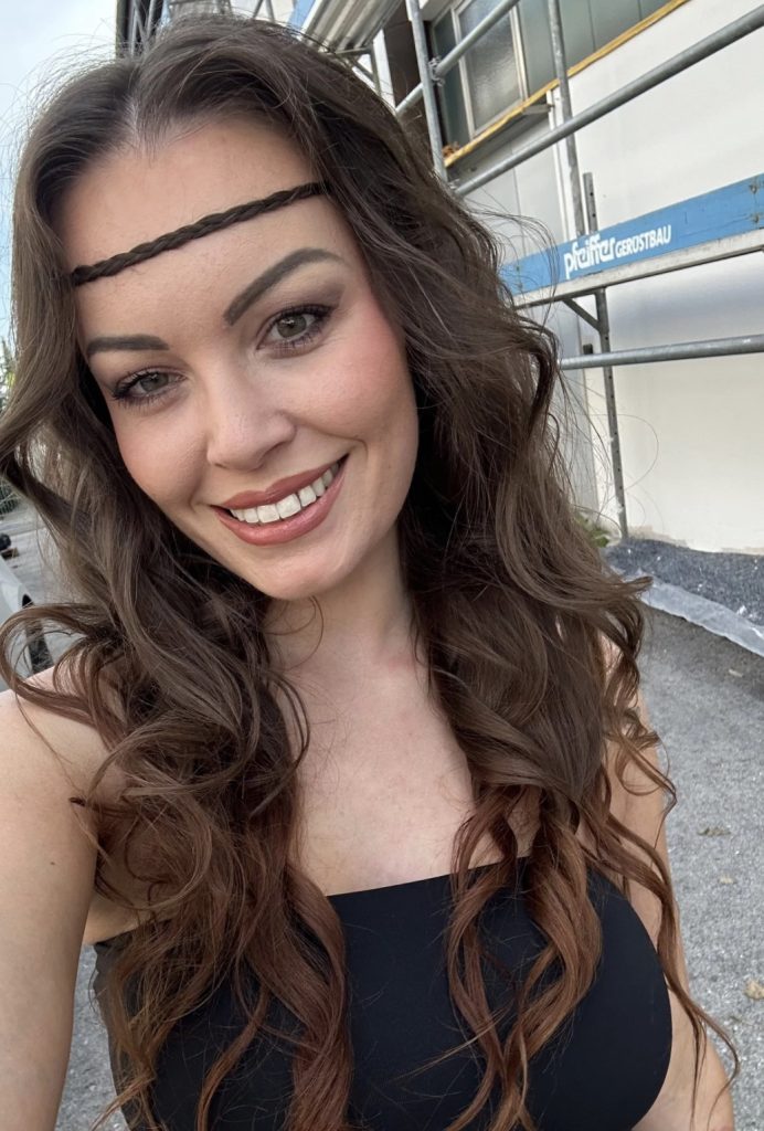 NanaSecret mit langen braunen Haaren, geflochtenem Haarband und schwarzem Oberteil bei einem Selfie im Freien.