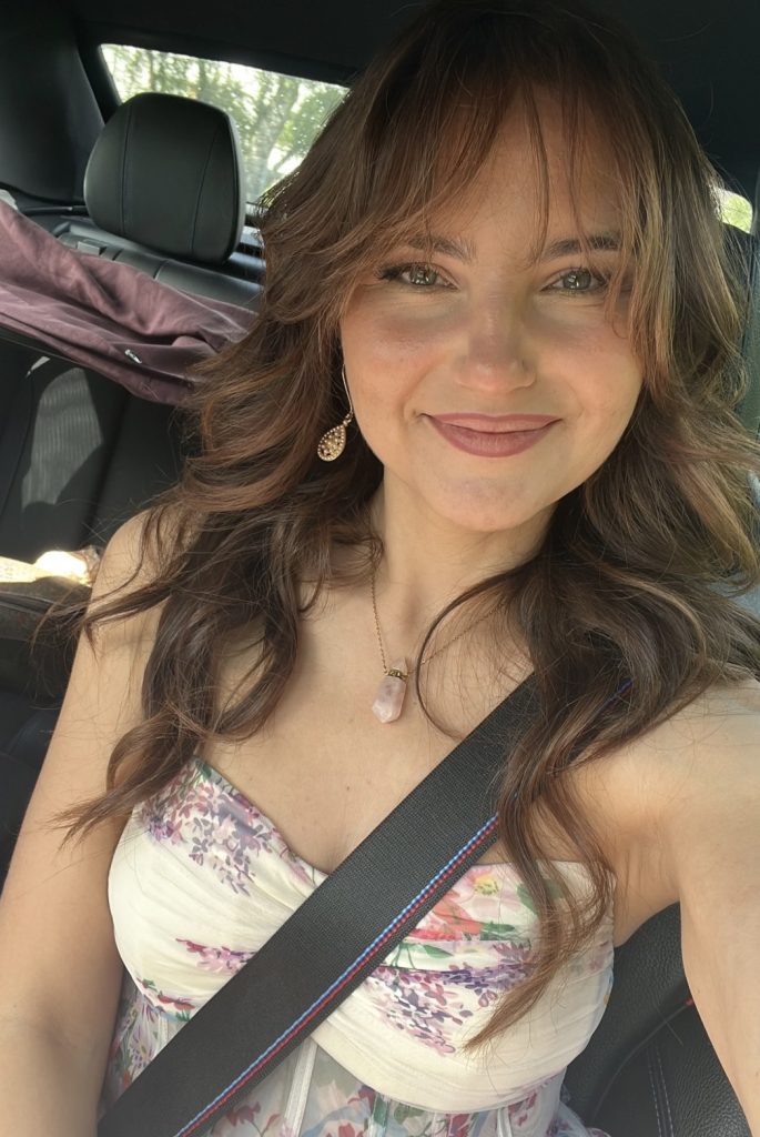 Mary X Ann mit langen braunen Haaren und floralem Outfit bei einem Selfie im Auto.