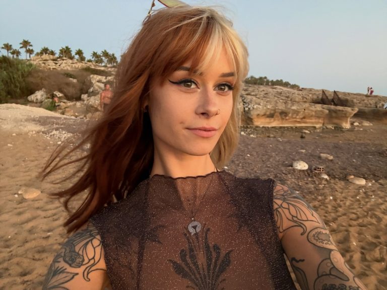 SallyDinosaur takes a natural selfie on the beach, showing her tattooed arms.