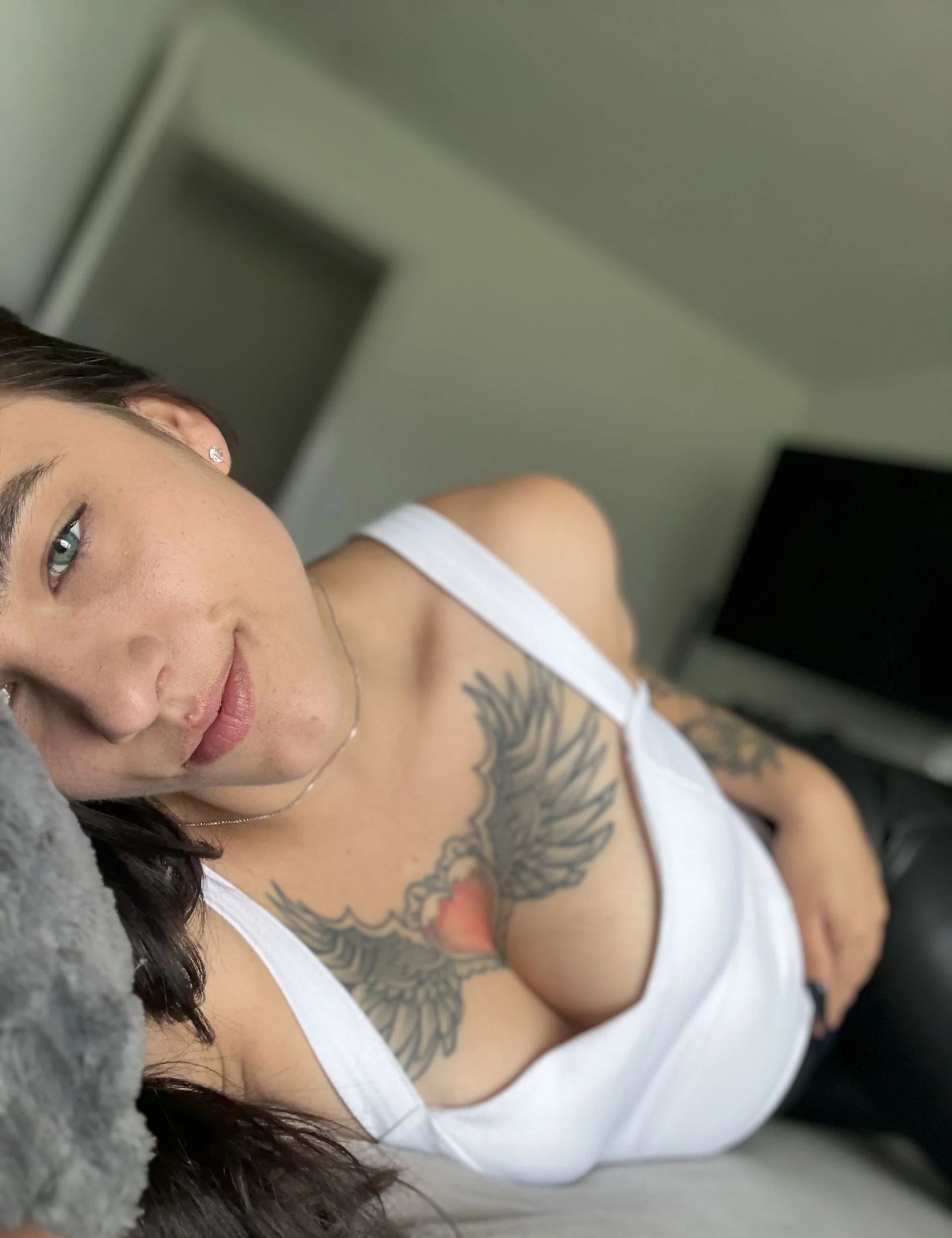Luna Lynn lies on a sofa in a white top and looks directly into the camera; her large heart-and-wings tattoo is clearly visible.