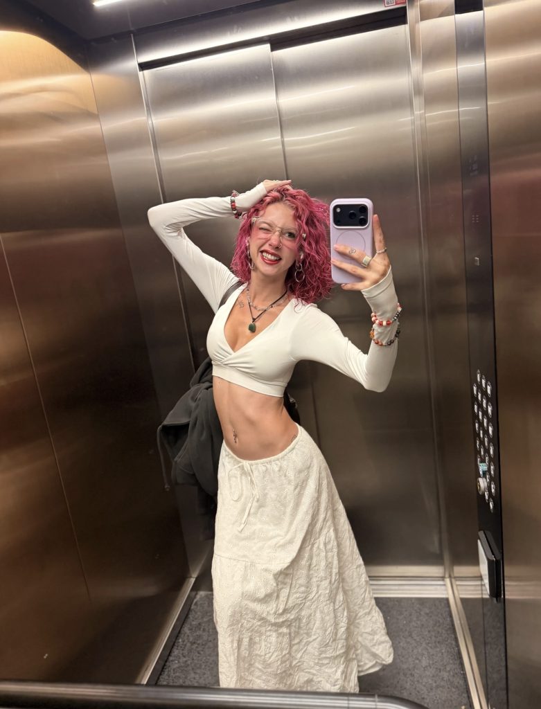 Melanie Jasmin mirror selfie in an elevator wearing a white crop top and maxi skirt